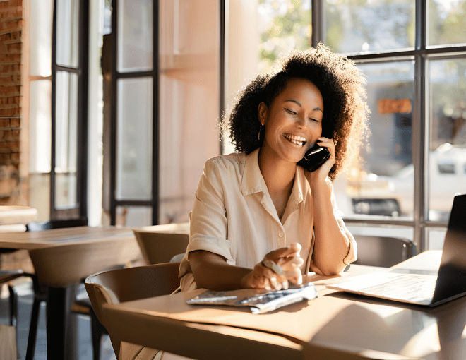 A woman seated at a cafe table, focused on her laptop, pauses to answer a phone call with a smile, sunlight filtering through the window behind her.